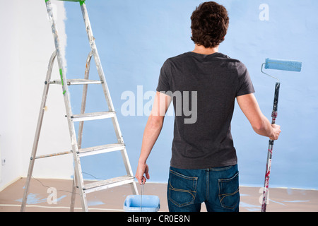 Young man holding a paint roller stands pensively in front of a wall Stock Photo