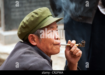 A mature man squat with a green hat smoking a pipe, Zhaoxing Dong Village, Southern China Stock Photo