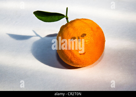 Blemishes and water drops altered to resemble a face on an orange with stem and a leaf on a textured light-colored background. Stock Photo