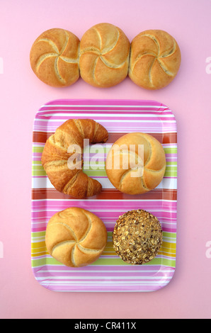 Bread rolls beside a croissant, a bread roll, a wholemeal roll and a multigrain roll on a striped, pink tray Stock Photo