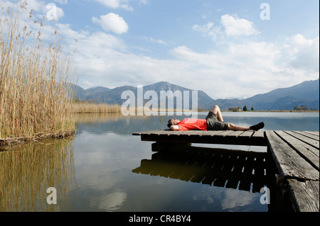 Man lying on a jetty on Eichsee lake in Kochler Moor near Schlehdorf Kochel, with Herzogstand und Jochberg mountains at back Stock Photo