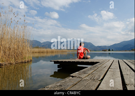 Man sitting on a jetty on Eichsee lake in Kochler Moor near Schlehdorf Kochel, with Herzogstand und Jochberg mountains at back Stock Photo