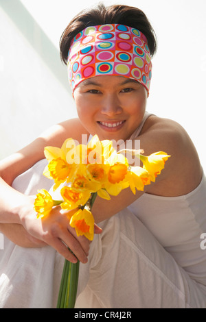 Portrait of healthy looking asian lady holding flowers wearing sleeveless white exercise outfit and smiling Stock Photo