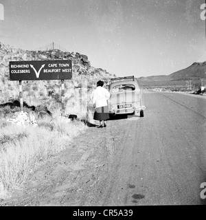 South Africa,1950s. A lady standing behind her small car parked on a dusty empty road in the outback of the vast African country, looking at a road or direction sign, showing arrows to different locations, including Cape Town, Beaufort West, Richmond and Colesberg. Stock Photo