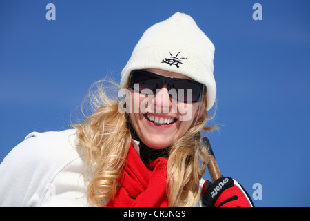 Young woman, about 25 years, wearing a bright cap, dark sunglasses and a red scarf, against a blue sky, holding ski sticks Stock Photo