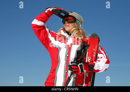 Young woman, about 25 years, wearing a red and white ski suit and a light grey cap, against a blue sky Stock Photo