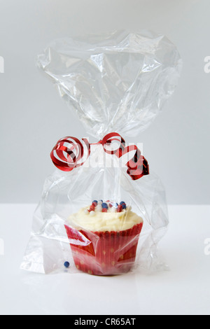 Cupcake in a colourful red wrapper and cellophane bag tied with a red ribbon against a white background Stock Photo
