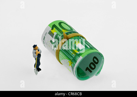 Miniature figure of a workman standing in front of roll of 100-euro notes and looking thoughtfully up Stock Photo