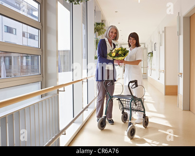 Germany, Cologne, Senior women and caretaker holding bouquet and standing by wheeled walker, smiling Stock Photo