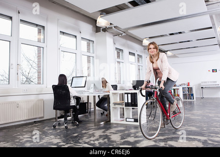 Germany, Bavaria, Munich, Woman cycling in office while colleagues working Stock Photo