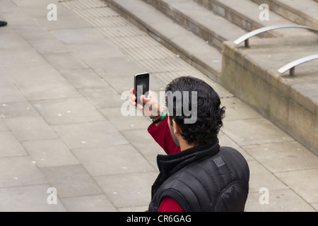 Young Asian man taking  photographs in a paved area of Manchester city centre using a camera built into a mobile phone. Stock Photo