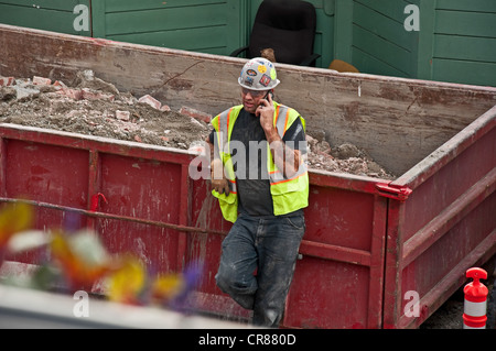This construction worker man is taking a break, leaning on a dumpster, while talking on a cell phone. Stock Photo
