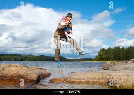 Man carrying a baby in his arms jumping across water, Mikkeli, Finland, Europe Stock Photo
