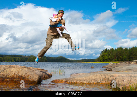 Man carrying a baby in his arms jumping across water, Mikkeli, Finland, Europe Stock Photo