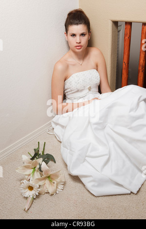 Nervous young bride wearing a wedding gown, sitting on an interior floor with her bouquet beside her. Stock Photo