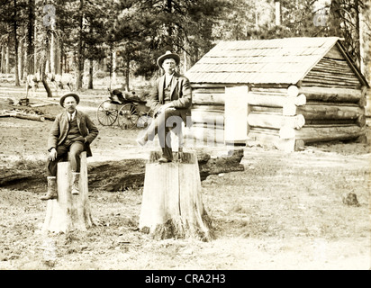 Two Men Sit on Treestumps in front of Small Log Cabin Stock Photo