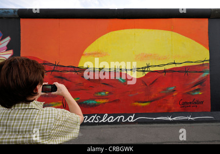 Tourist taking a photograph of Art and the Berlin Wall, sunrise in the East behind barbed wire, painting on a remaining segment Stock Photo