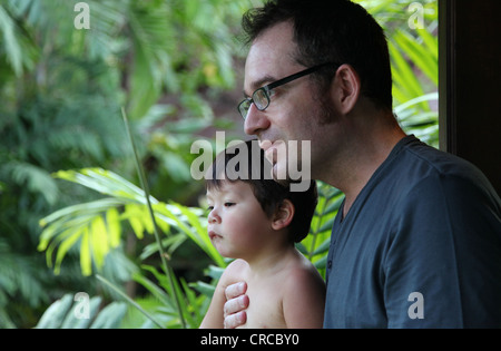 It's a photo of a baby boy and his dad. They are at the balcony of a bungalow hotel looking at the jungle. Dad Wear glasses. Sun Stock Photo