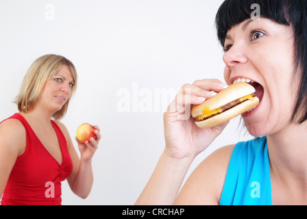 two women eating burger and apple, with the blond woman with apple in the background, looking enviously at the woman with the burger Stock Photo