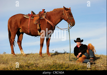 Cowboy sitting on the ground, holding the horse by its reins, looking into the distance, Saskatchewan, Canada, North America Stock Photo