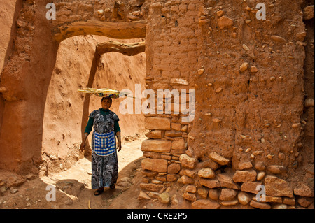 Elderly Berber woman walking through an alleyway in a clay village, Ksar, carrying a bowl of twigs on her head, Tamdakht Stock Photo