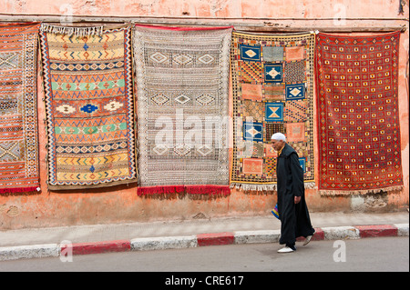 Man wearing a headdress, a traditional Djellabah, and slippers, Babouche, walking past a house wall with carpets hanging for Stock Photo