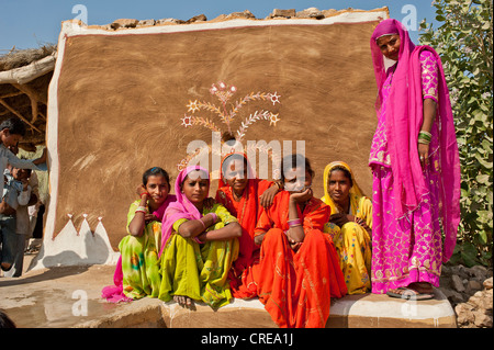 Young girls wearing brightly colored saris attending a wedding, they are sitting on the floor in front of a wall painted with Stock Photo
