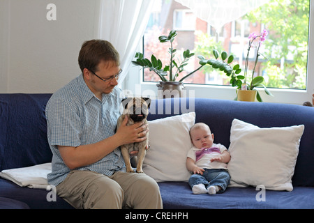 Pug (Canis lupus f. familiaris), man with male Pug and three month old baby on sofa in living room Stock Photo