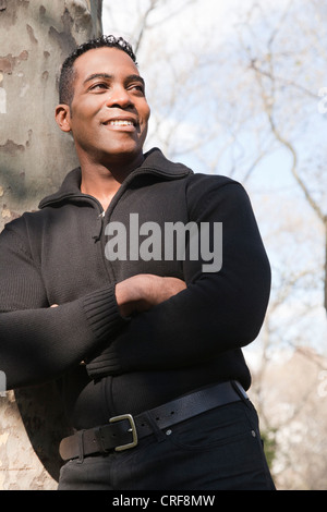 Smiling African American man with arms crossed, leaning on tree trunk Stock Photo