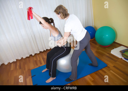 young woman doing exercises with theraband sitting on exercise ball, instructed by physio therapist Stock Photo