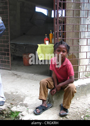 Young boy sitting in front of garage in which he sells locally produced alcohol to bypassers, Haiti, Province de l'Ouest, Kenskoff, Port-Au-Prince Stock Photo