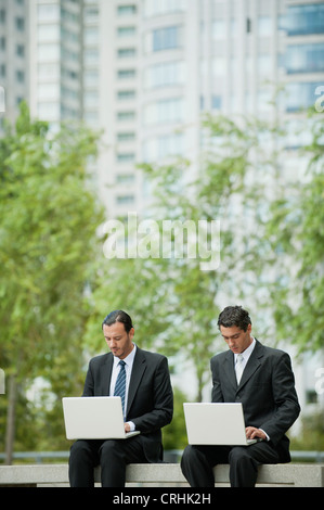 Businessmen sitting side by side using laptop computers outdoors Stock Photo