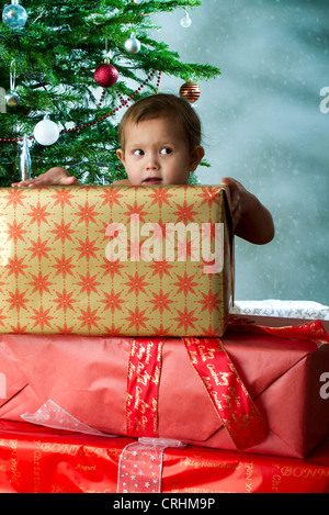 Baby girl standing behind stack of Christmas gifts with mischievous expression Stock Photo