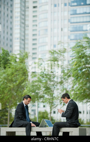Businessmen sitting outdoors with laptop computers, one man text messaging with cell phone Stock Photo