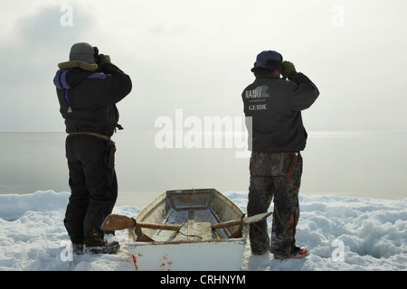 two Inuit sealers standing at the ice edge beside their rowing boat and look out on the open water with binoculars, Greenland, Ostgroenland, Tunu, Kalaallit Nunaat, Scoresbysund, Kangertittivag, Kap Tobin, Ittoqqortoormiit Stock Photo