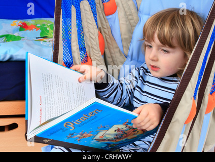 little boy in pyjama reading a children's book about pirates in a swing of a Billi-Bolli loft bed Stock Photo