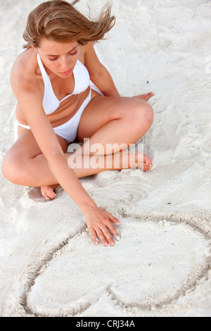 beautiful young bikini woman sitting at the beach, drawing a heart in the sand Stock Photo