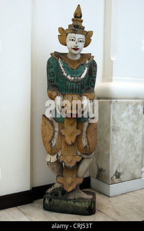 It's a photo of a Thai wood statue that stands near a white wall in the lobby of a building. It's painted ans starts to crack Stock Photo