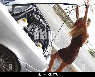 stumped young blond woman looking under the engine bonnet of her broken down car Stock Photo
