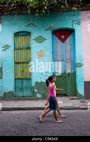 Two girls walking down the street in Old Havana Cuba, with a Cuban flag and viva Cuba libre painted on two doors in background. Stock Photo