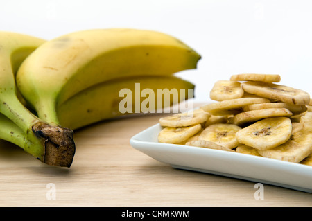 Dried banana chips in small white bowl on wooden chopping board against a white background with bunch of fresh bananas also Stock Photo
