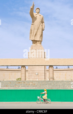 With motion blur.  A young Uyghur Chinese girl on on a bicycle travels past the statue of Mao Zedong in Kashgar. Stock Photo