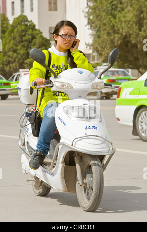 A Chinese woman travels along a road on her scooter while talking on her mobile phone in Kashgar. Stock Photo