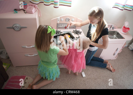 Two older sisters playing with one year old sibling in pretend play kitchen at home. Stock Photo