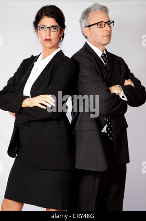 Cute, mature business couple, dressed in suits and wearing glasses, standing back to back with their arms crossed Stock Photo