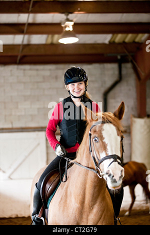 Teenage girl on horseback wearing helmet and safety vest in indoor arena Stock Photo