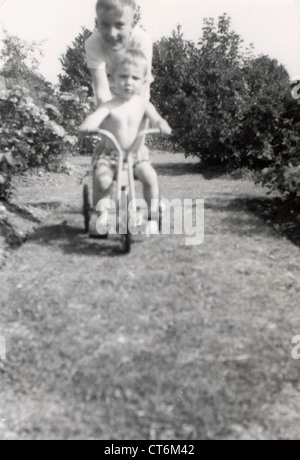 black and white photo a young boy pushing a younger boy on his tricycle outside in the garden family album Stock Photo