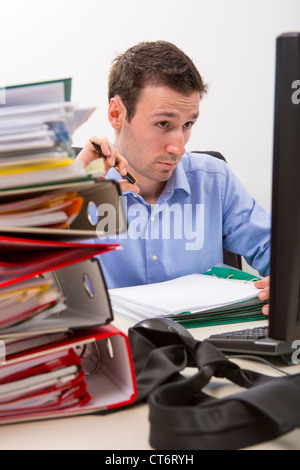 Confused accountant looking at some info on the display of his computer, surrounded by huge piles of documents. Stock Photo