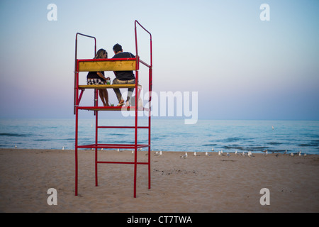 A young couple sitting on a lifeguard chair with a nearby lifeboat on ...