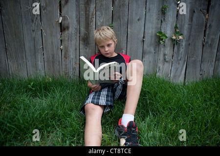 Ten year old boy reading a book, sitting in the grass and leaning against fence. Stock Photo